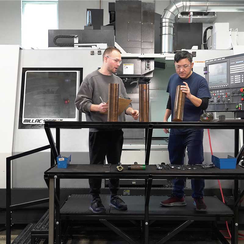 Engineers working with metal components near a computer numerical control (CNC) machine in a manufacturing facility.