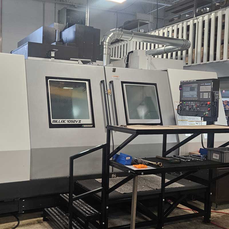 Industrial CNC milling machine (MILLAC 1052VII) with closed doors and control panel in a manufacturing shop, featuring metal ducting overhead and a work table with tools in the foreground.