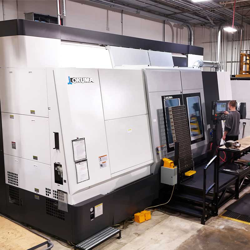 Large Okuma CNC machining center on a factory floor with an operator using the control panel during a manufacturing process.